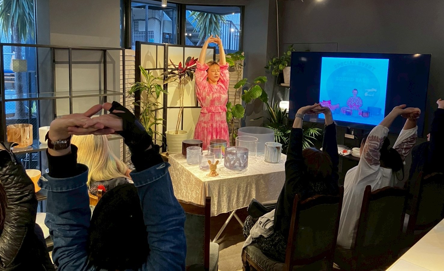 ピンクの服の春名めぐみが腕を高く伸ばし、参加者もいっしょに身体を伸ばしている、クリスタルボウルのイベント
